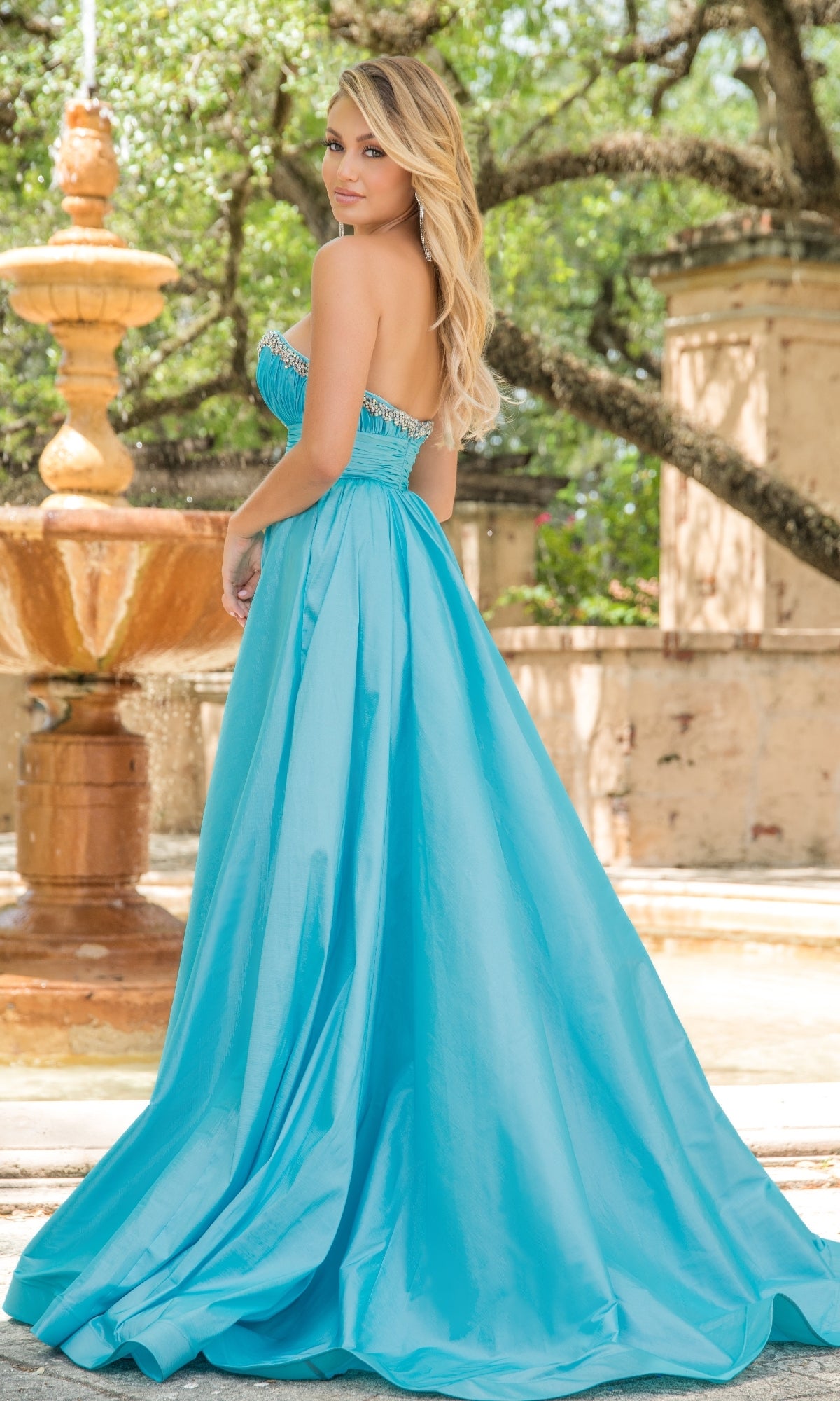 A woman in the Long Formal Dress 38337 by Ava Presley, a strapless light blue A-line gown, stands outdoors near a stone fountain with trees and sunlight in the background.